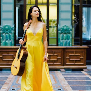 Anabel Montesinos i gul kjole går gjennom et lyst rom med gitaren i hånda.