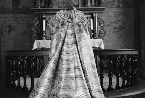 Liv Kristin Holmberg bakfra med fotsid kappe foran et alter, i svarthvitt.