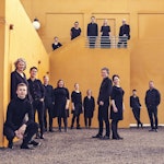 Sangerne i Det Norske Solistkor står i et orange rom, i svarte klær, spredt rundt på gulv og oppå en balkong.