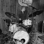 Svart-hvitt-bilde av Isak brekken som spiller trommer foran et skilt der det står «Pleti – kulturbanken».