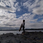 Mar Bonet står med celloen ved strandkanten foran fjorden. Det er hvite skyer på blå himmel.