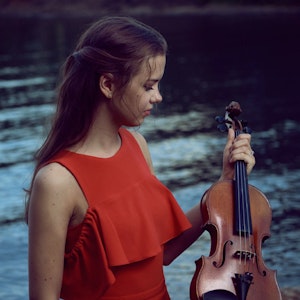 Fiolinist Maren Ohm Ballestad i rød kjole foran en fjord og med fiolinen i hånda.