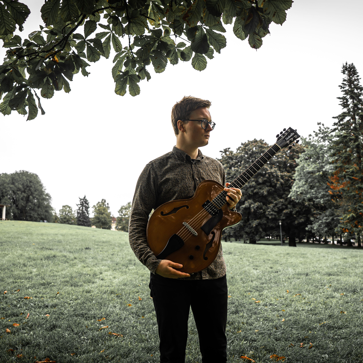 Markus Kaardal står på en slette under et tre med gitar i hendene.