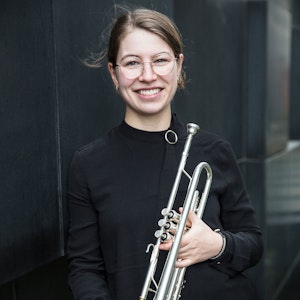 Monika Holst Olsen står foran mørk vegg og smiler med trompeten i hånda.