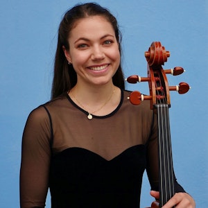 Sophia Hugo Cabo står foran blå vegg og smiler med celloen i hånden.