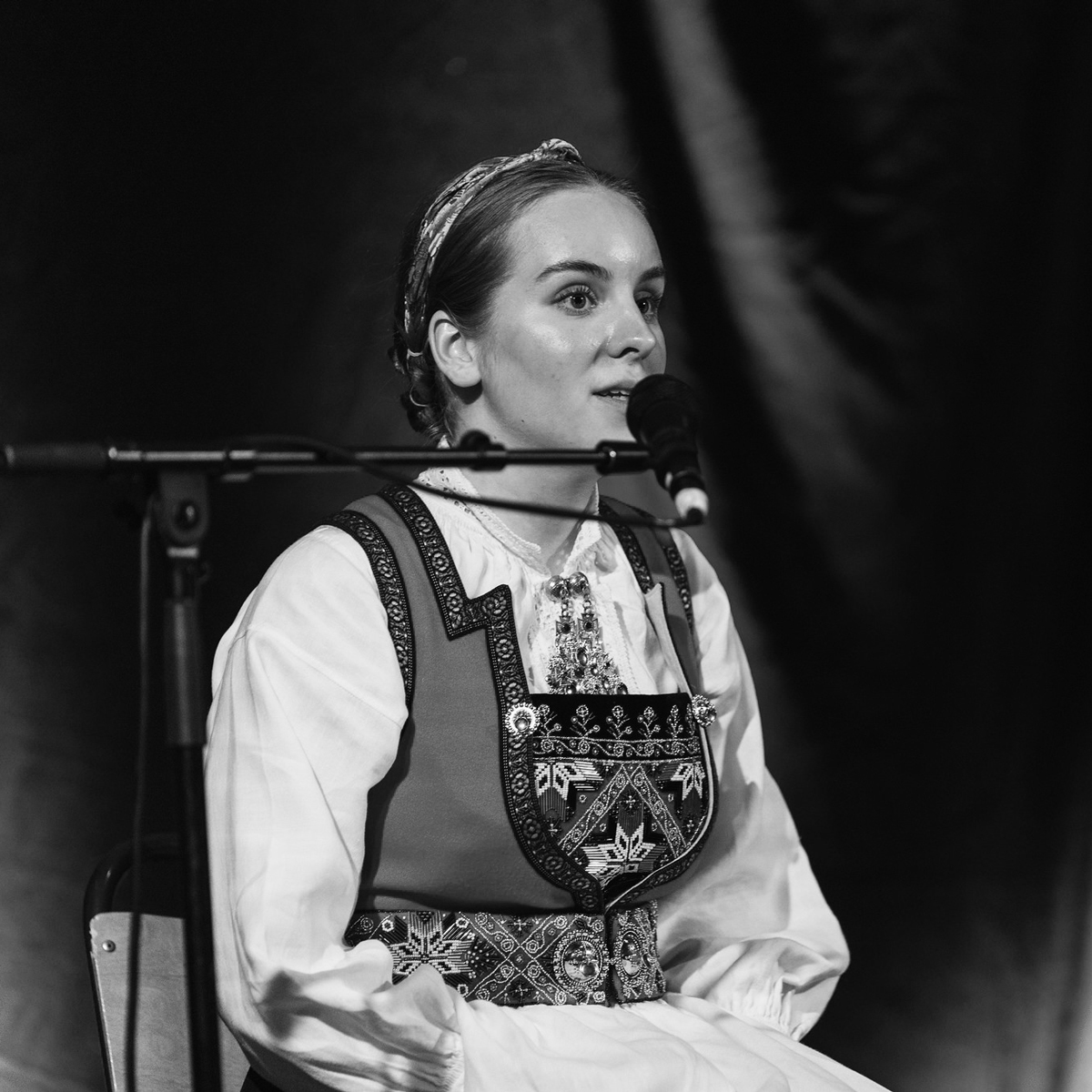 Anna Russnes sitter iført bunad og synger i en mikrofon.