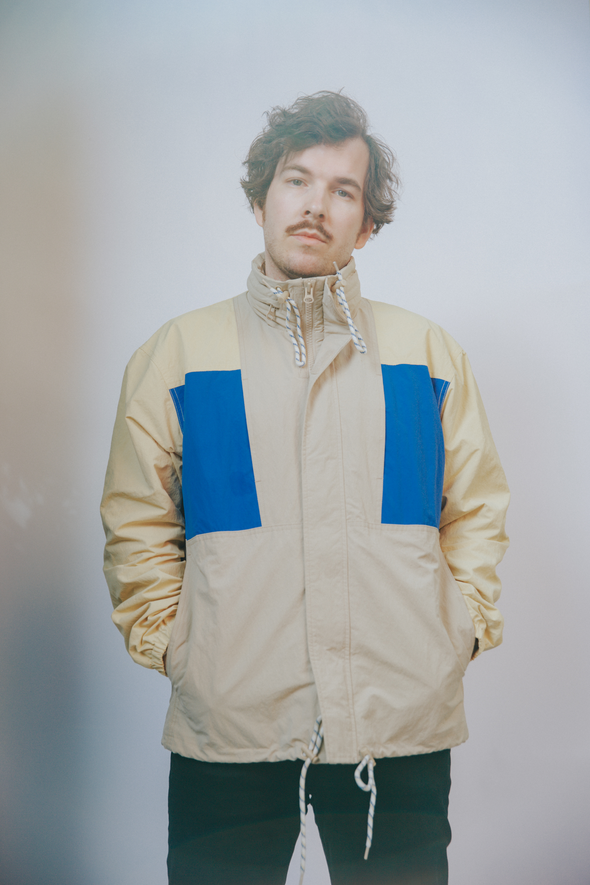 Berque står i beige og blått med henden i lommene foran hvit vegg.