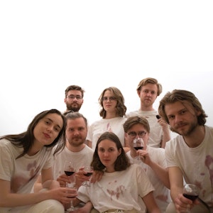 Medlemmene i bandet LEAH sitter i band-t-skjorter i en gruppe foran kritthvit bakgrunn.