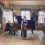 Lotte Hellstrøm Hestad i herskapelig sal. Hun står på scenen med fiolinen med pianist bak seg.