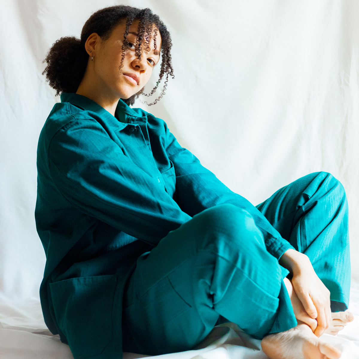 Sanyu Nsubuga sitter i lotusstilling og turkise klær foran hvit bakgrunn.