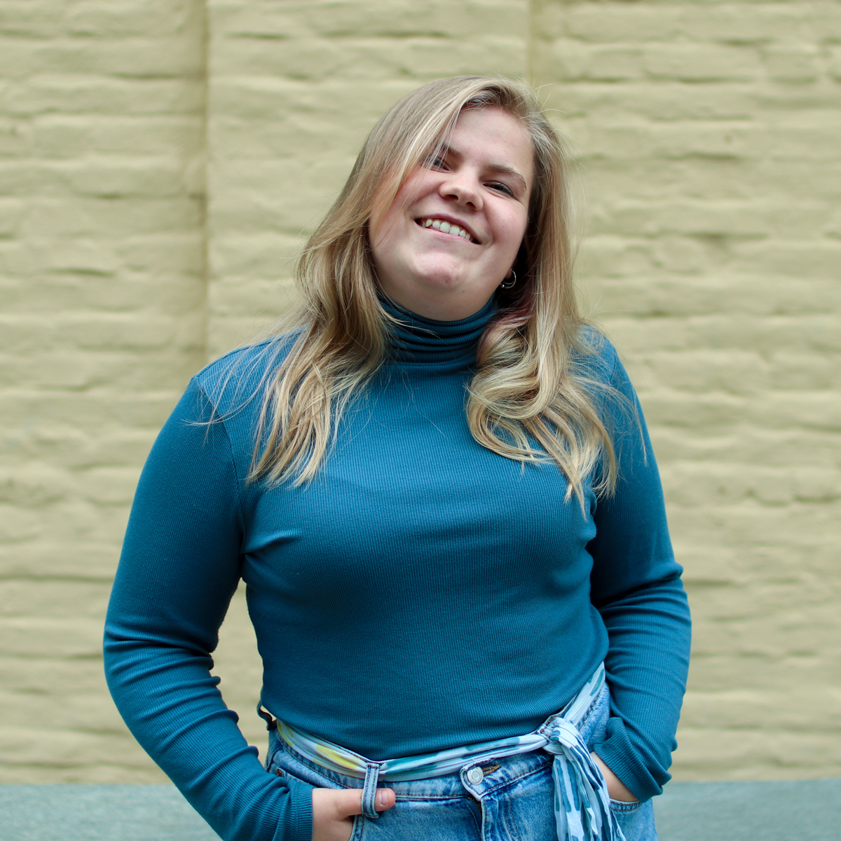 Sigyn Sjølie Sandegren står foran gulgrønn vegg med hendene i lommene og smiler.