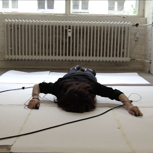 Parkins lies on the floor with her face down towards a large sheet of paper. Wires are attached around both wrists.