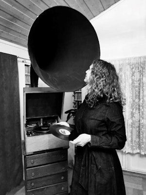 A girl is standing in a room holding a LP and looking into the speaker of a gramophone player.