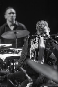Kalle is playing on the accordion. He is placed on a crowded stage between two drum sets.