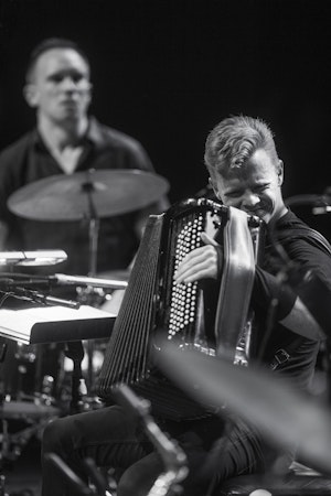 Kalle is playing on the accordion. He is placed on a crowded stage between two drum sets.