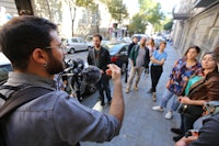 En man står vendt mot en foklemengde i en sør-Europeisk gate. Han holder et filmkamera og gestikulerer noe med den frie hånden.