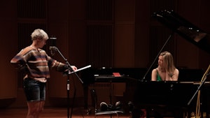 Live Maria Roggen stands upright in front of a microphone and points toward sheet music. Ingfrid Breie Nyhus is sitting behind a grand piano while looking at Live.