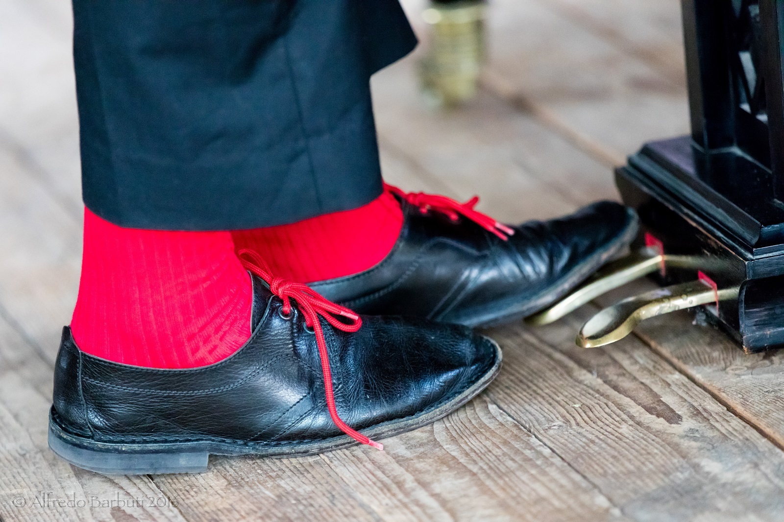 Nærbilde av Misha Alperins føtter, med illrøde sokker og sorte sko, og hvor venstre fot trykker på pedalen på et flygel.