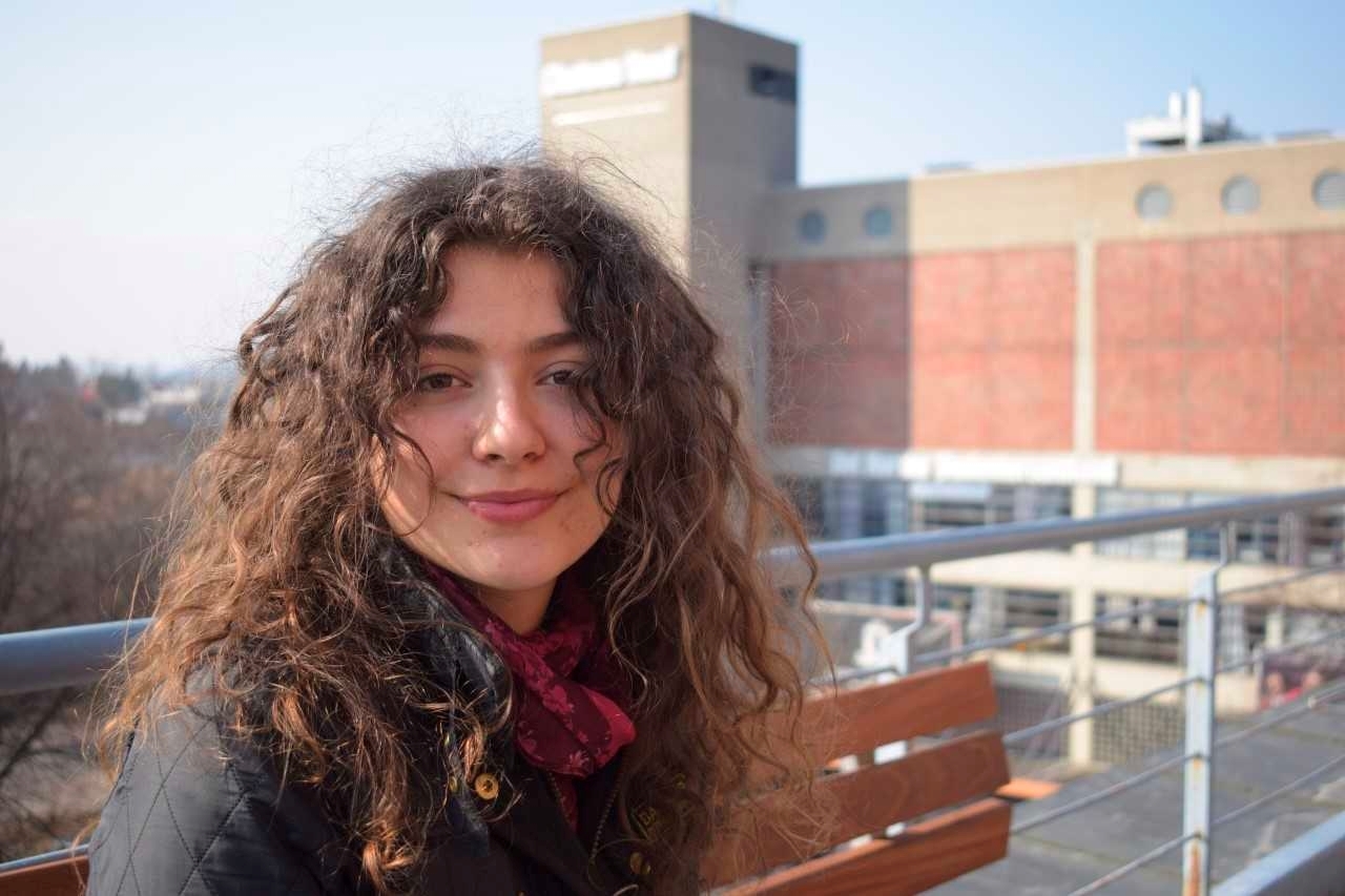 Idun sitter på en benk på terassen, med Chateau Neuf i bakgrunnen. Hun har langt, krøllete hår og smiler mot kamera.