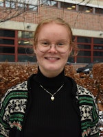 Karen Elise stands outside in front of a snow-covered bush and smiles at the camera. She has glasses, turtleneck and a woolen cardigan.
