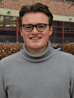 Sigmund stands in front of a snow-covered bush and smiles at the camera. He is wearing glasses and a turtleneck jumper.