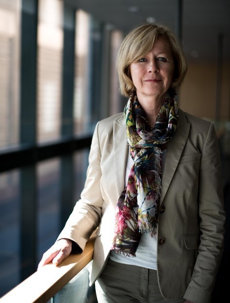 Ingrid står ved store vinduer, holder i et rekkverk og smiler forsiktig mot kameraet.