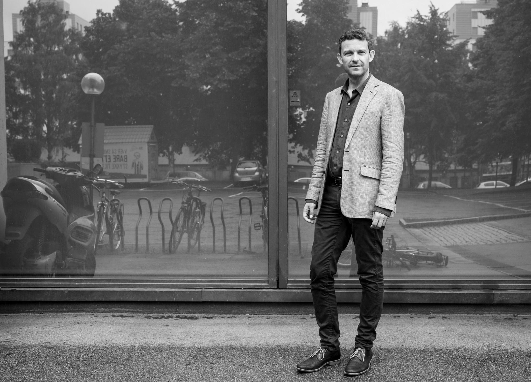 Jon Helge står utenfor Musikkhøgskolen foran et av vinduene i første etasje. Gjenskinnet viser en park, trær, sykkelstativ og en moped.