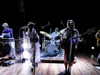 Et band på fem står på scenen og spiller – en på trommer, en på bass, en på gitar, en med dataen som instrument og en som synge.