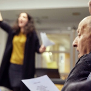 Dirigent som løfter armen og en eldre person som synger fra en tekst