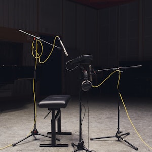 Mørkt lydstudio. Opptaksutstyr, pianokrakk, mikrofoner på stativ. Mikrofonene har ledninger i knall gult.