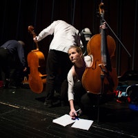 Tre studenter står på en scene, to med celloer og en vendt mot publikum skriblende på noteark.