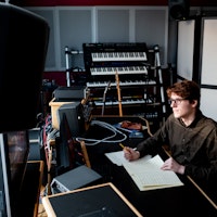 En student befinner seg i et studio proppfull av synther og høyttalere. Han sitter ved en pult og noterer ned noter på et ark.