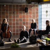 Fem komponiststudenter sitter på gulvet foran en vegg hvor én holder en cello og én taster på en datamaskin. Lys skinner inn gjennom et vindu.