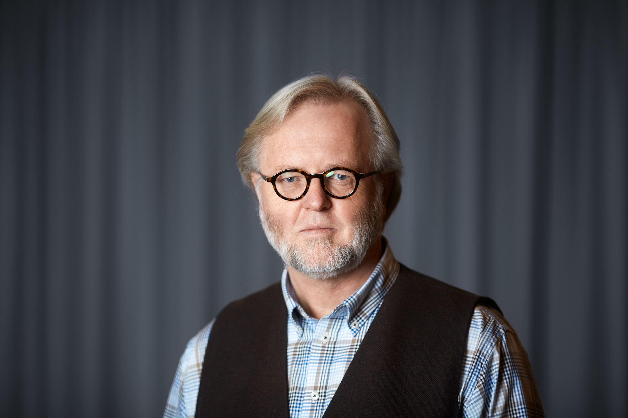 Øivind står foran en mørk vegg og ser alvorlig mot kamerat. På seg har han runde briller, skjorte og vest.